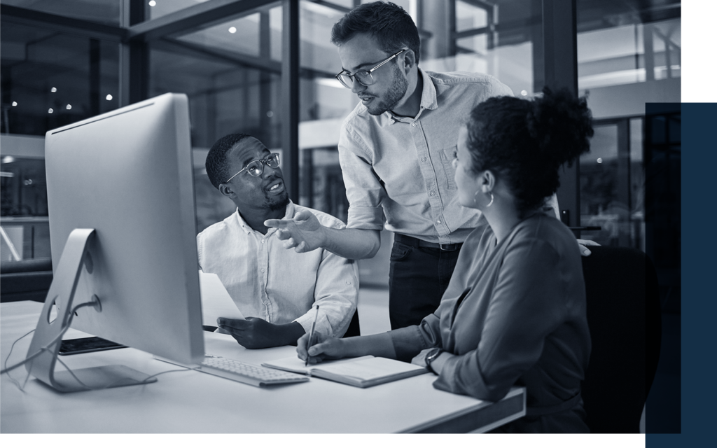 Mix of standing and seated team members engaged in discussion while looking at shared monitor on table